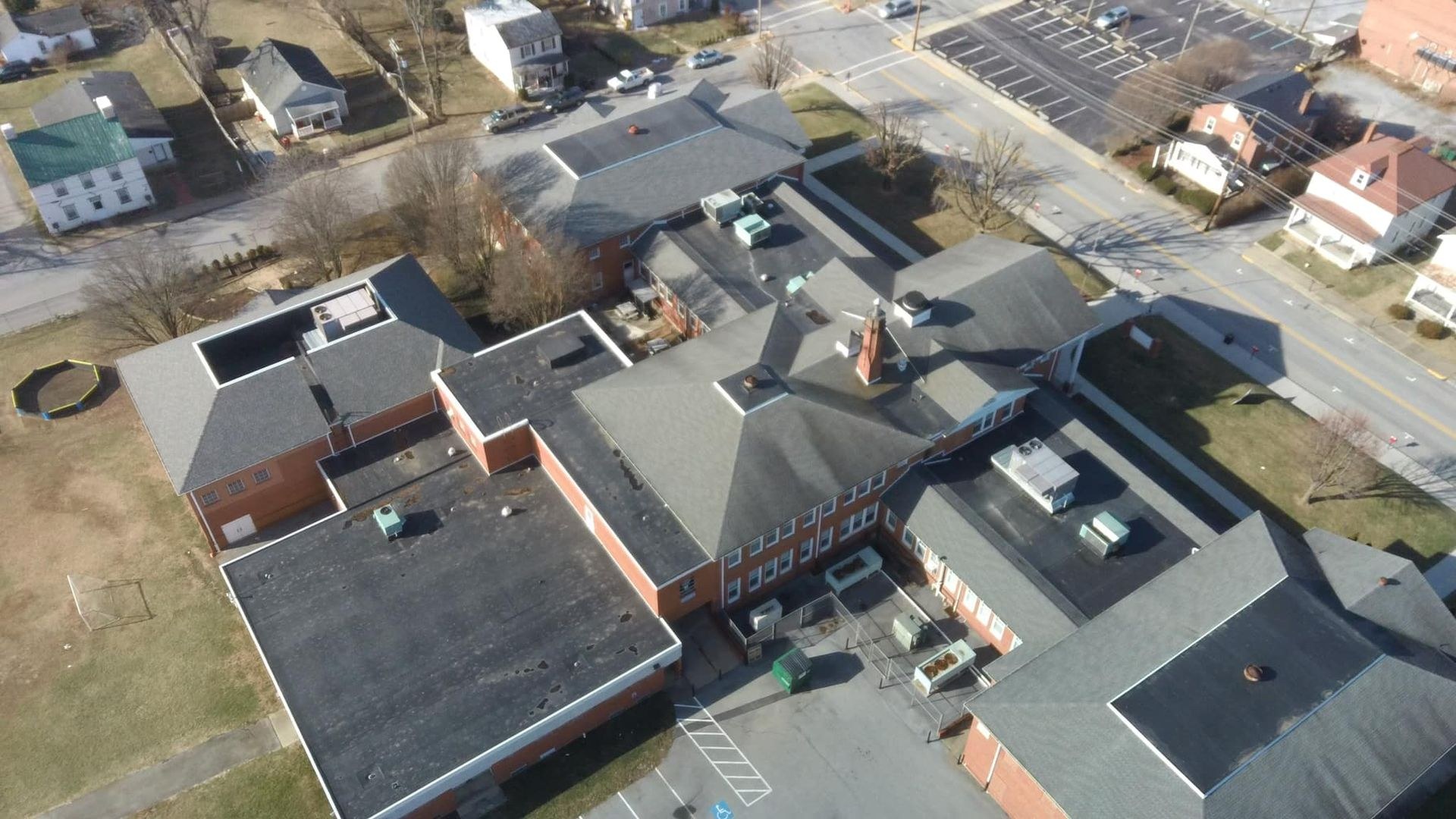 Aerial drone view of a large brick school building in Martinsburg WV showing gray asphalt shingle roof sections alongside low slope flat membrane roof areas with rooftop HVAC units