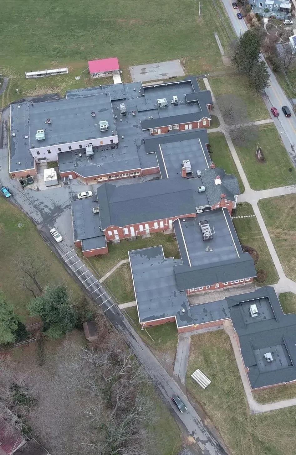Aerial drone view of a brick school campus in the Martinsburg WV area showing large low slope commercial roof sections with rooftop HVAC units alongside steep shingled roof wings