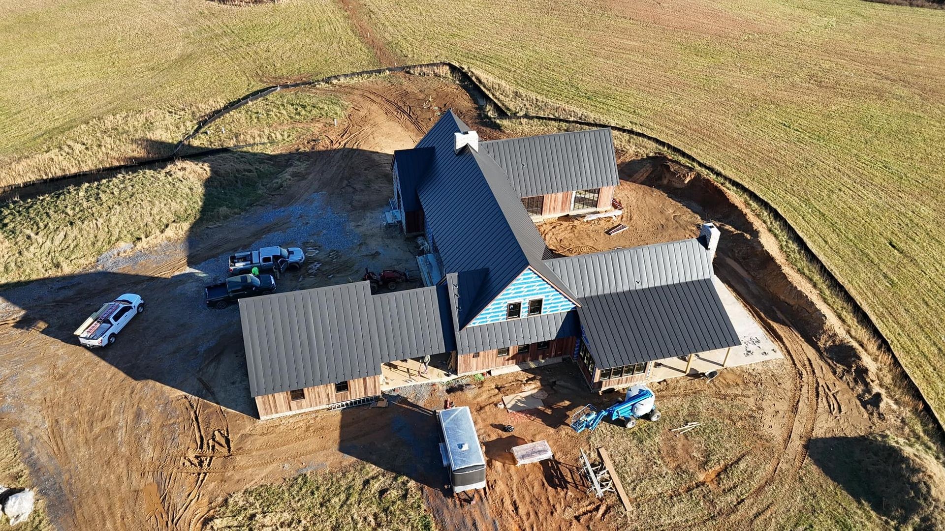 Aerial view of a new custom home with a dark charcoal standing seam metal roof installed by Black Stone Roofing near Martinsburg WV, with board and batten siding still going up and work trucks parked on the jobsite