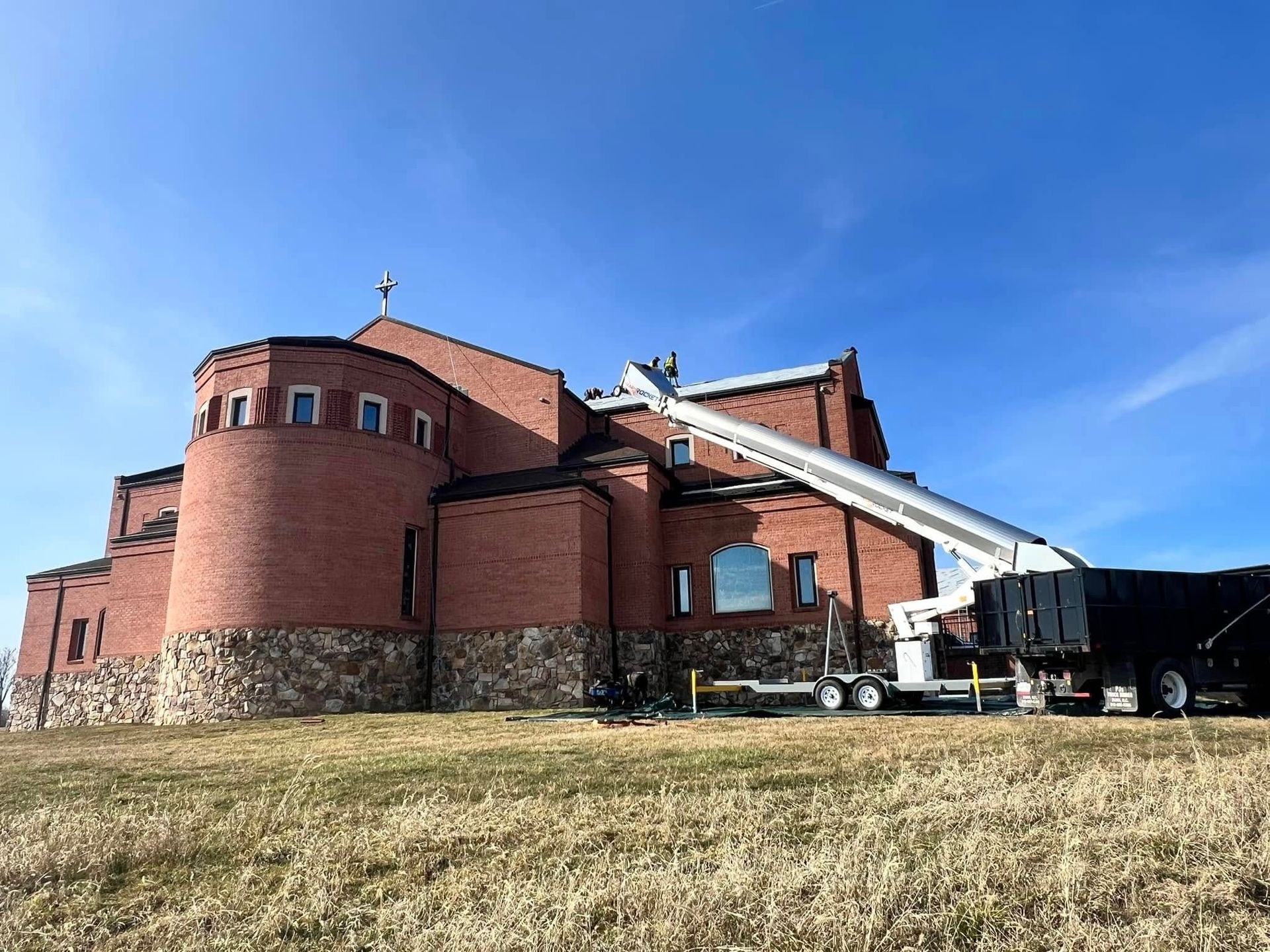 Roofing crew working on a large brick church in Martinsburg WV with a truck mounted conveyor boom lifting material up to the roof line and a debris trailer staged on the lawn below