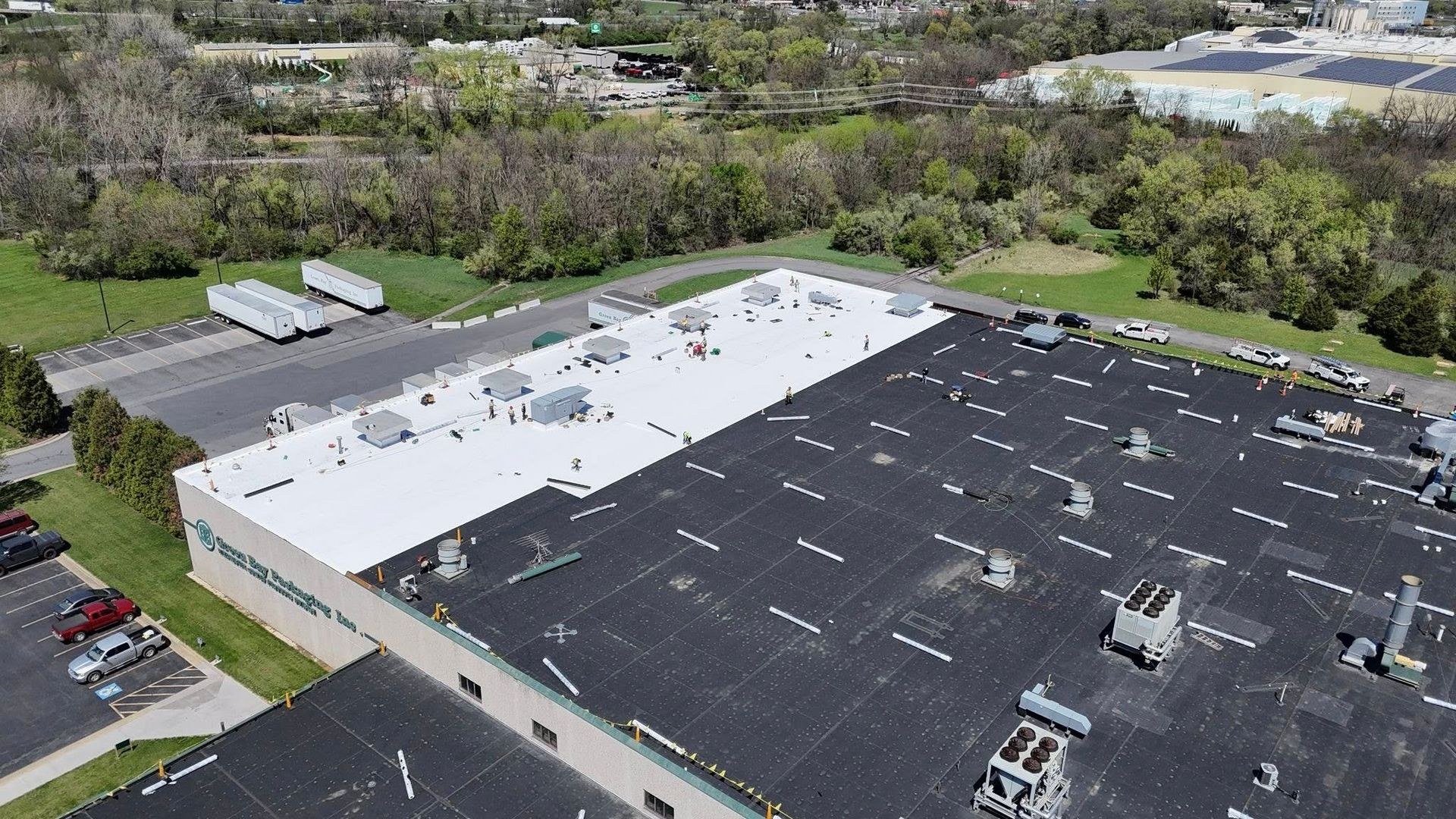 Aerial view of a commercial reroof in progress where new white single ply membrane covers part of a large packaging plant roof and the original black roof remains on the rest, crew working the transition line for Black Stone Roofing of Martinsburg WV