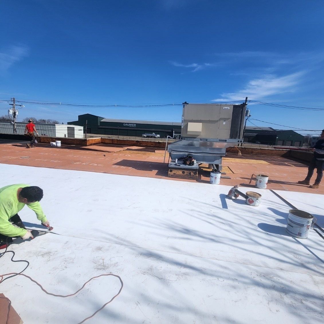 Roofer in green high visibility gear kneeling to run a seam tool along freshly installed white single ply membrane beside a rooftop HVAC unit and buckets of adhesive on a flat commercial roof, crew at work for Black Stone Roofing of Martinsburg WV