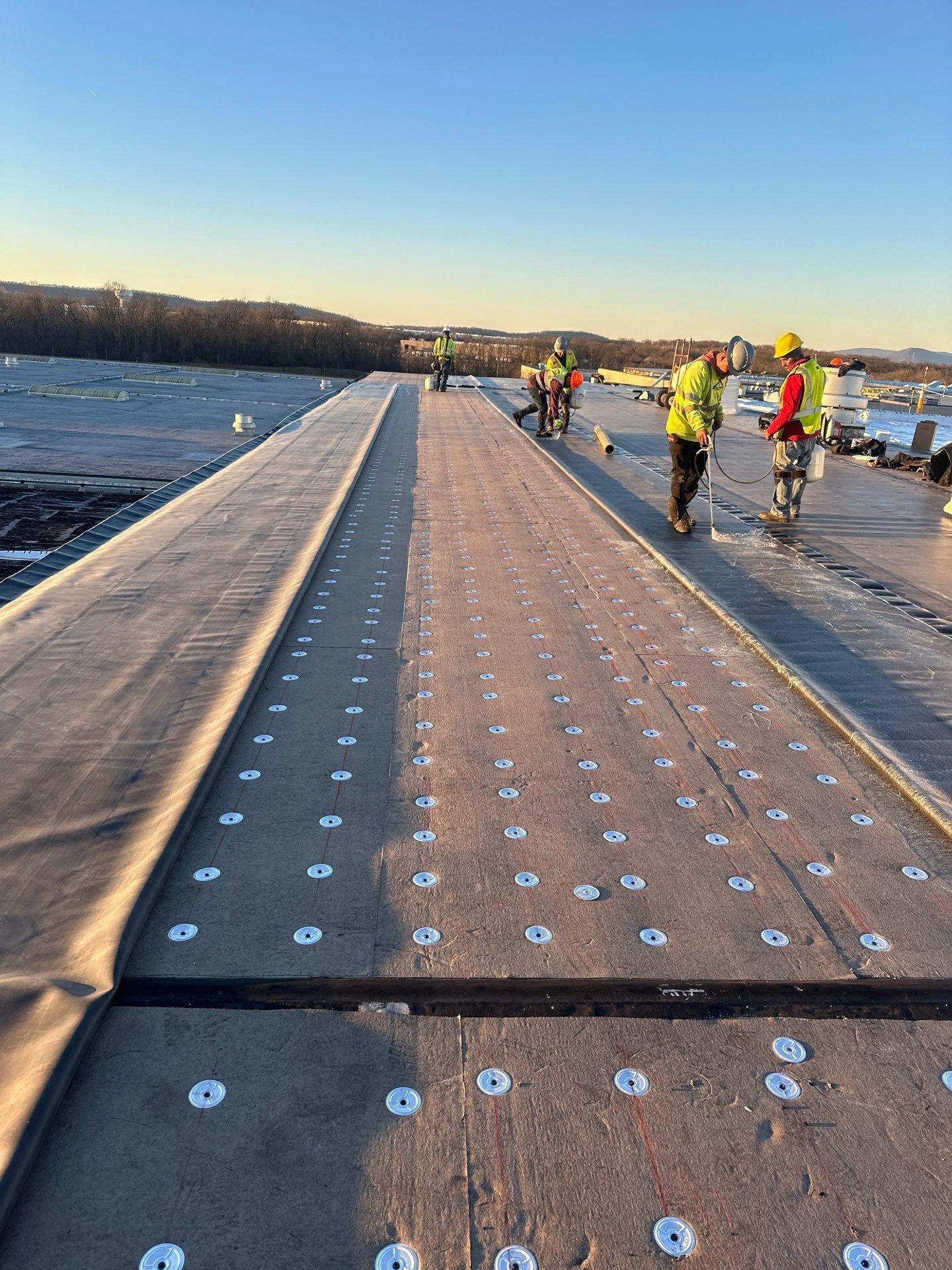 Roofing crew installing mechanically fastened single ply membrane on a large commercial flat roof in Martinsburg WV with rows of white fastener plates across the seams and skylights on the adjacent metal roof section