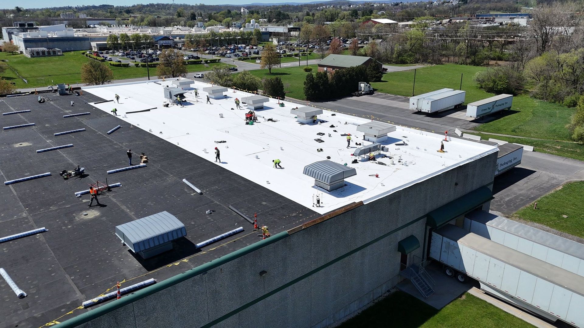 Aerial view of a roofing crew installing white single ply membrane across a warehouse roof with wrapped membrane rolls staged in rows on the remaining black roof section, commercial roofing work by the Black Stone Roofing team based in Martinsburg WV