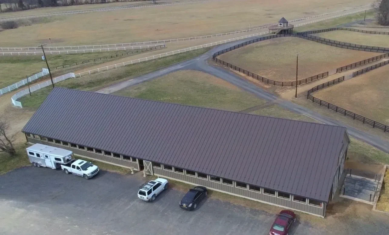 Aerial view of a long horse barn near Martinsburg WV with a brown standing seam metal roof, surrounded by fenced pastures and a gravel lot with a truck and horse trailer